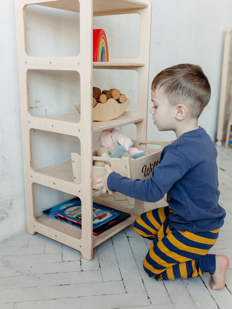 May include: A light wood bookshelf with four shelves and a drawer. The drawer is labeled "Willie". A child is kneeling and opening the drawer. The bookshelf is filled with toys, books, and other items.