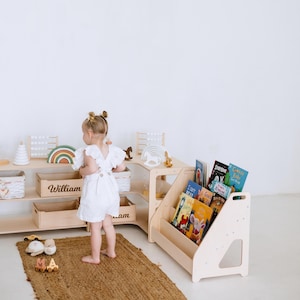 May include: A child's playroom with a wooden bookshelf filled with colorful books. A wooden storage unit with open shelves and a wooden toy rainbow. A small child in a white romper stands on a jute rug.