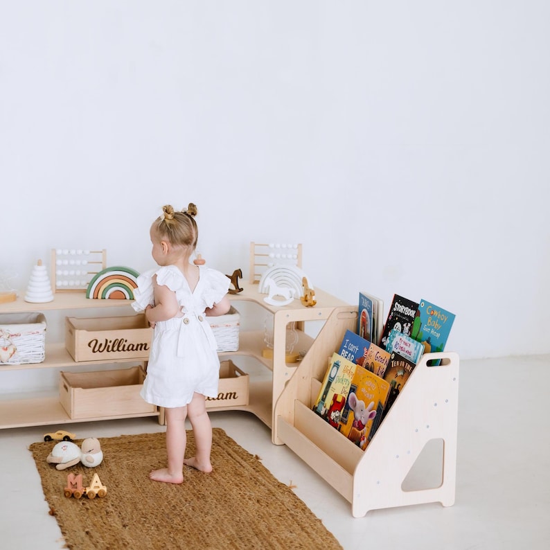 May include: A child's playroom with a wooden bookshelf filled with colorful books. A wooden storage unit with open shelves and a wooden toy rainbow. A small child in a white romper stands on a jute rug.