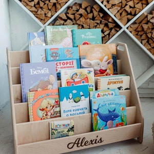 May include: A wooden bookshelf with five shelves, filled with colorful children's books. The bookshelf is labeled "Alexis" at the bottom.