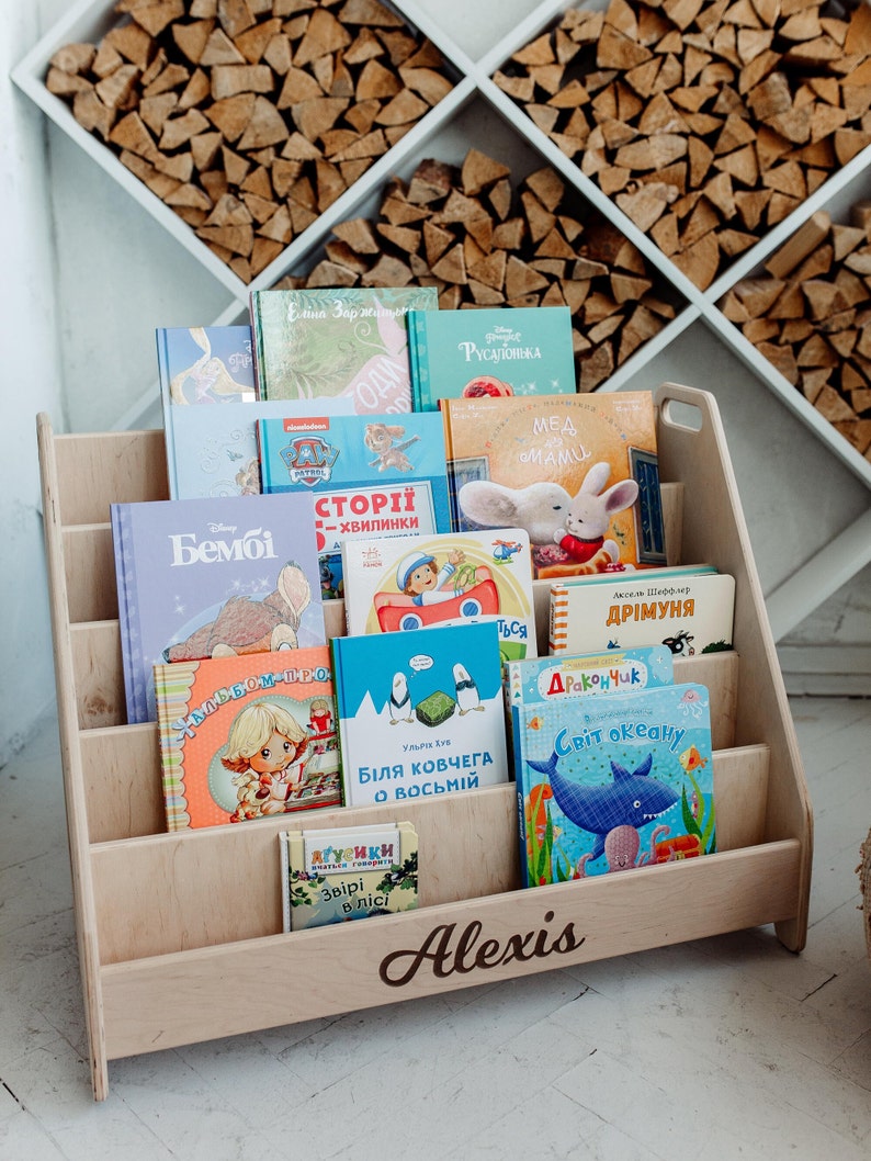 May include: A wooden bookshelf with five shelves, filled with colorful children's books. The bookshelf is labeled "Alexis" at the bottom.
