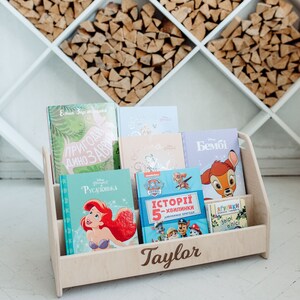 May include: A wooden bookshelf with the name "Taylor" engraved on the front. The bookshelf is filled with children's books, including "The Little Mermaid", "Paw Patrol", and "Bambi".