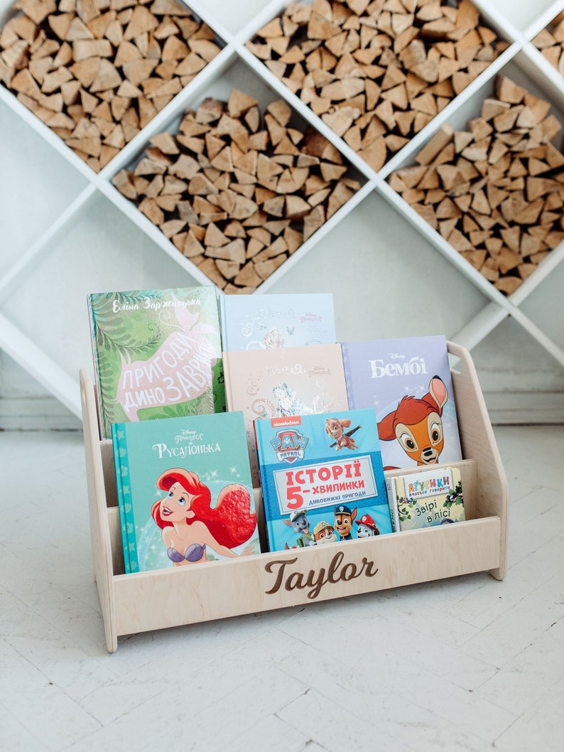 May include: A wooden bookshelf with the name "Taylor" engraved on the front. The bookshelf is filled with children's books, including "The Little Mermaid", "Paw Patrol", and "Bambi".