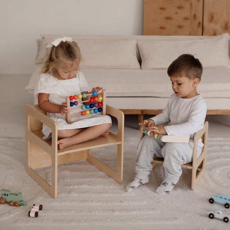 May include: Two children are seated on small, light-colored wooden chairs, each holding a toy. One child has a colorful abacus, while the other has a wooden toy with pastel-colored beads. The setting is a bright room with a neutral-toned sofa and rug.
