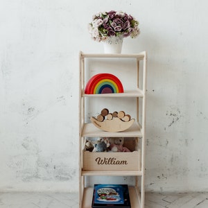 May include: A white wooden shelf unit with five shelves. The top shelf has a bouquet of purple and white flowers in a white pot. The second shelf has a rainbow-colored wooden toy. The third shelf has a wooden toy shaped like a crescent moon with wooden balls on top. The fourth shelf has a wooden box with the name "William" engraved on the front. The bottom shelf has a book with a blue cover.