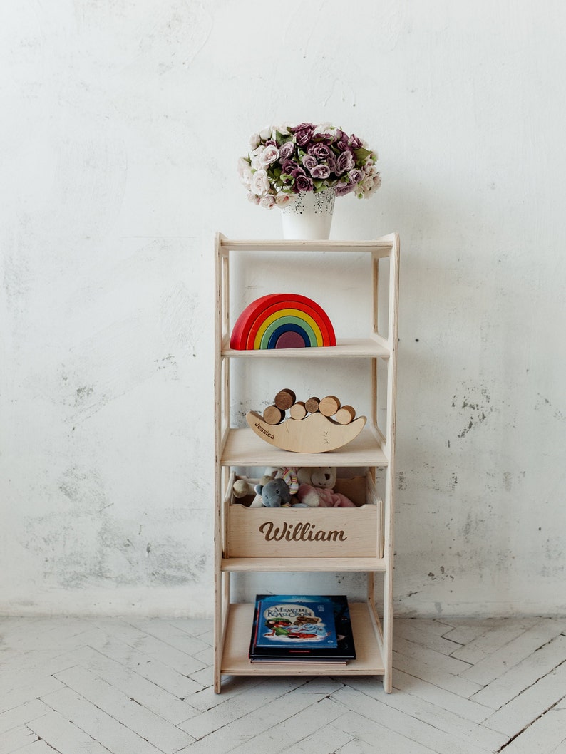 May include: A white wooden shelf unit with five shelves. The top shelf has a bouquet of purple and white flowers in a white pot. The second shelf has a rainbow-colored wooden toy. The third shelf has a wooden toy shaped like a crescent moon with wooden balls on top. The fourth shelf has a wooden box with the name "William" engraved on the front. The bottom shelf has a book with a blue cover.