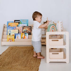 May include: A light wood bookshelf with a variety of children's books on the shelves. A child is standing next to a light wood play table with a rainbow arch and a wooden abacus. The play table has two shelves with storage bins. The child is wearing a white shirt and grey shorts. The play area is on a natural fiber rug.