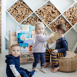 May include: Three children are playing with books in a room with a white wall and a wooden shelf filled with firewood. The shelf is labeled "Alex". The children are wearing blue, purple, and yellow clothing.