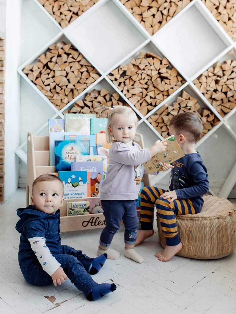 May include: Three children are playing with books in a room with a white wall and a wooden shelf filled with firewood. The shelf is labeled "Alex". The children are wearing blue, purple, and yellow clothing.
