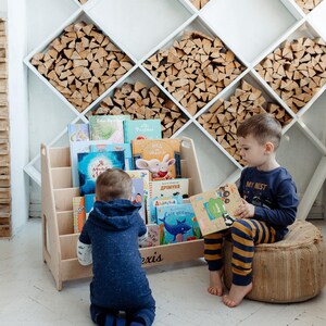 May include: Two children, one sitting on a woven cushion and the other kneeling on the floor, are reading books from a wooden bookshelf. The bookshelf has a variety of colorful children's books with Russian titles. The bookshelf is labeled "Alexis".