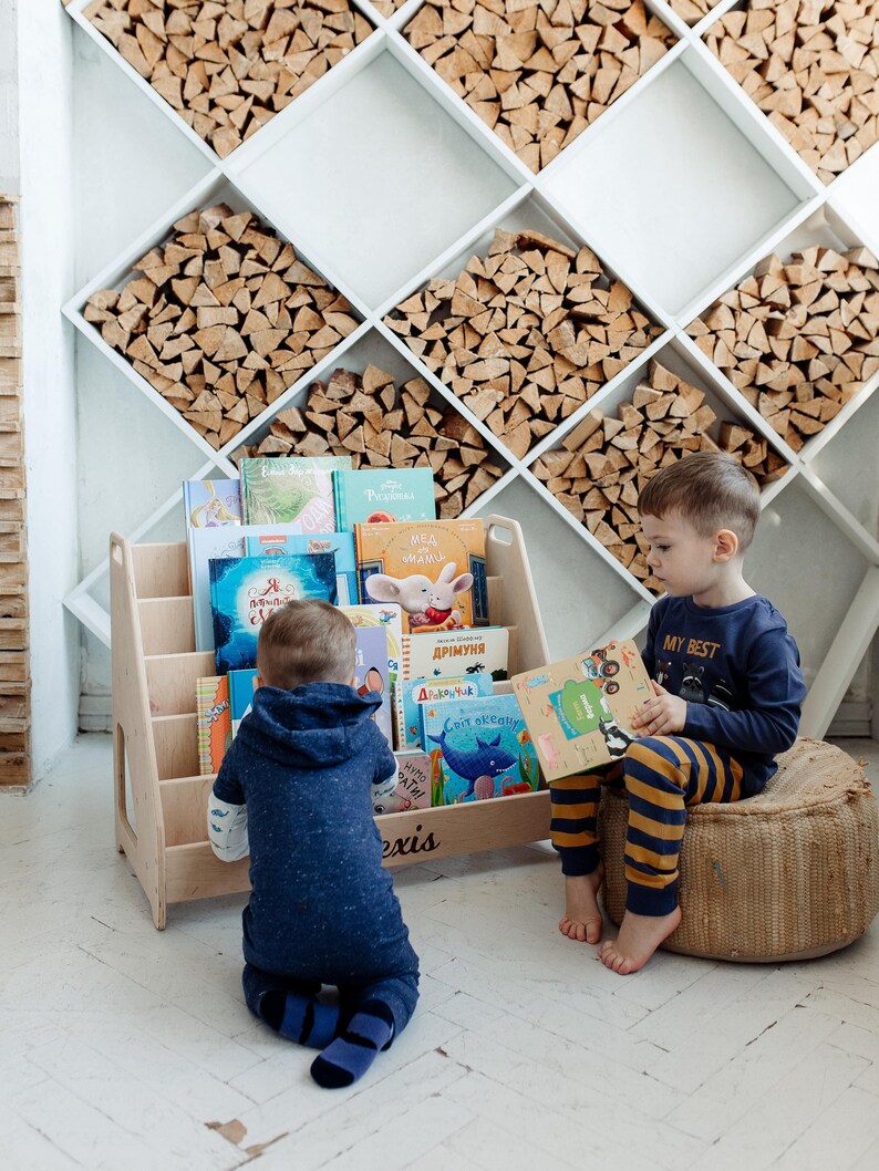 May include: Two children, one sitting on a woven cushion and the other kneeling on the floor, are reading books from a wooden bookshelf. The bookshelf has a variety of colorful children's books with Russian titles. The bookshelf is labeled "Alexis".
