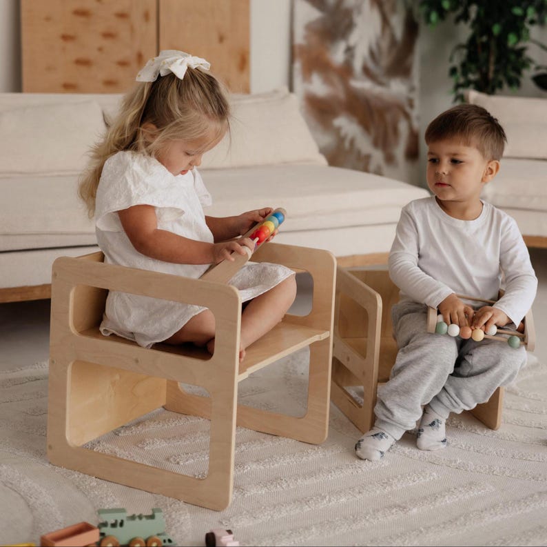 May include: Two small, light-colored wooden chairs. A child sits in each chair, one holding a colorful toy and the other holding a wooden toy with pastel-colored beads. The chairs have a simple, modern design, suitable for toddlers.
