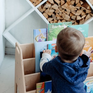 May include: A wooden bookshelf with multiple shelves filled with colorful children's books. A young child is reaching for a book on the top shelf.