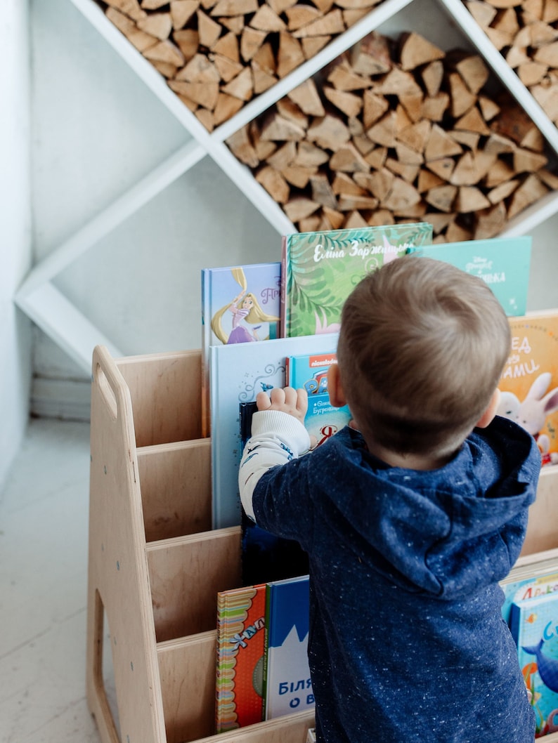 May include: A wooden bookshelf with multiple shelves filled with colorful children's books. A young child is reaching for a book on the top shelf.