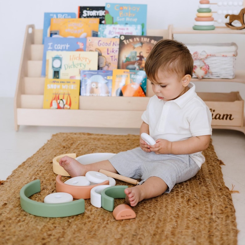 May include: A child sits on a woven rug surrounded by wooden toys. The toys include curved arches in green, white, and yellow, along with round and rectangular pieces. A bookshelf with children's books is in the background.