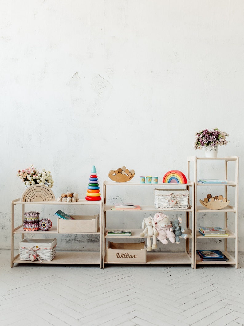May include: White wooden shelves with rainbow stacking rings, a rainbow arch, a wooden rainbow, a basket, books, stuffed animals, and a wooden box labeled "William".
