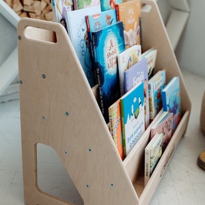 May include: A wooden bookshelf with a handle, designed for children, is filled with colorful books. The bookshelf is made of light wood and has a natural finish. The books are arranged in rows, with the titles facing outward. The bookshelf is a great way to encourage children to read and learn.