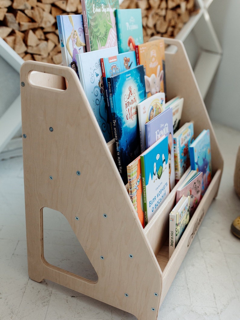 May include: A wooden bookshelf with a handle, designed for children, is filled with colorful books. The bookshelf is made of light wood and has a natural finish. The books are arranged in rows, with the titles facing outward. The bookshelf is a great way to encourage children to read and learn.