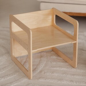 May include: A small, light-colored wooden chair with a simple design. The chair has a seat, back, and armrests with cut-out sections. The chair is made of light-colored wood and is likely for a child.