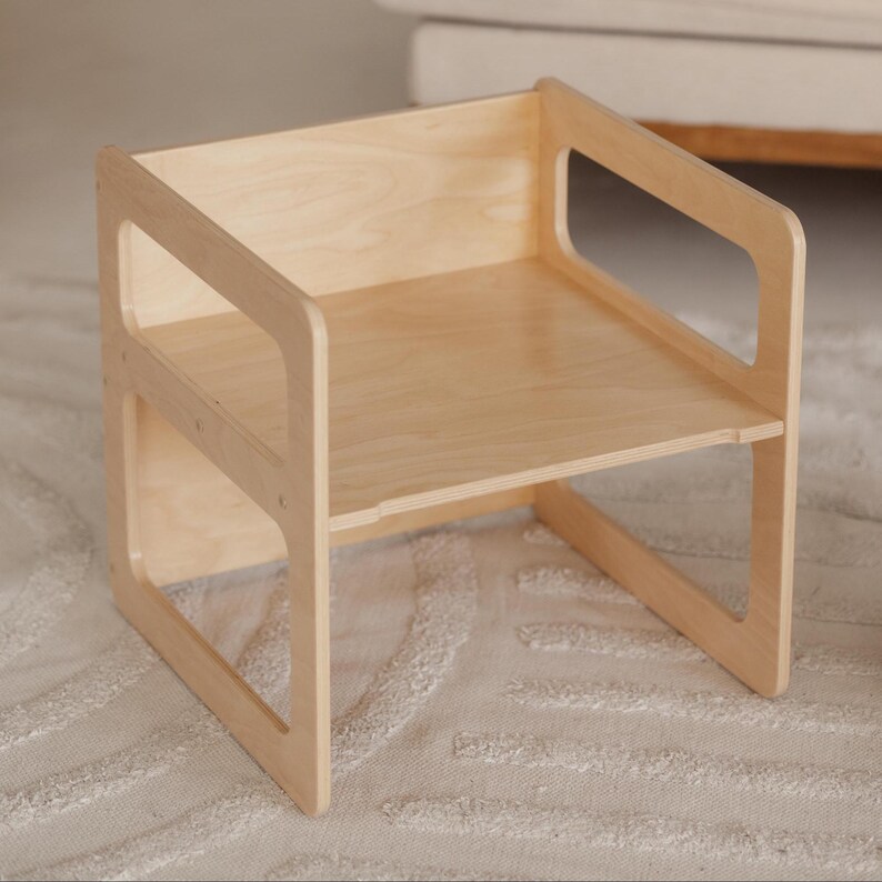 May include: A small, light-colored wooden chair with a simple design. The chair has a seat, back, and armrests with cut-out sections. The chair is made of light-colored wood and is likely for a child.