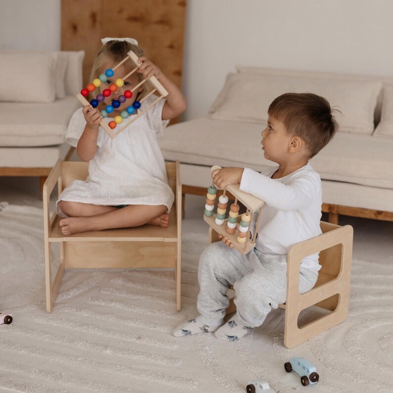 May include: Two children are seated on small wooden chairs, each holding a wooden abacus toy with colorful beads. The abacuses have rainbow-colored beads and pastel-colored beads. The chairs are made of light-colored wood.