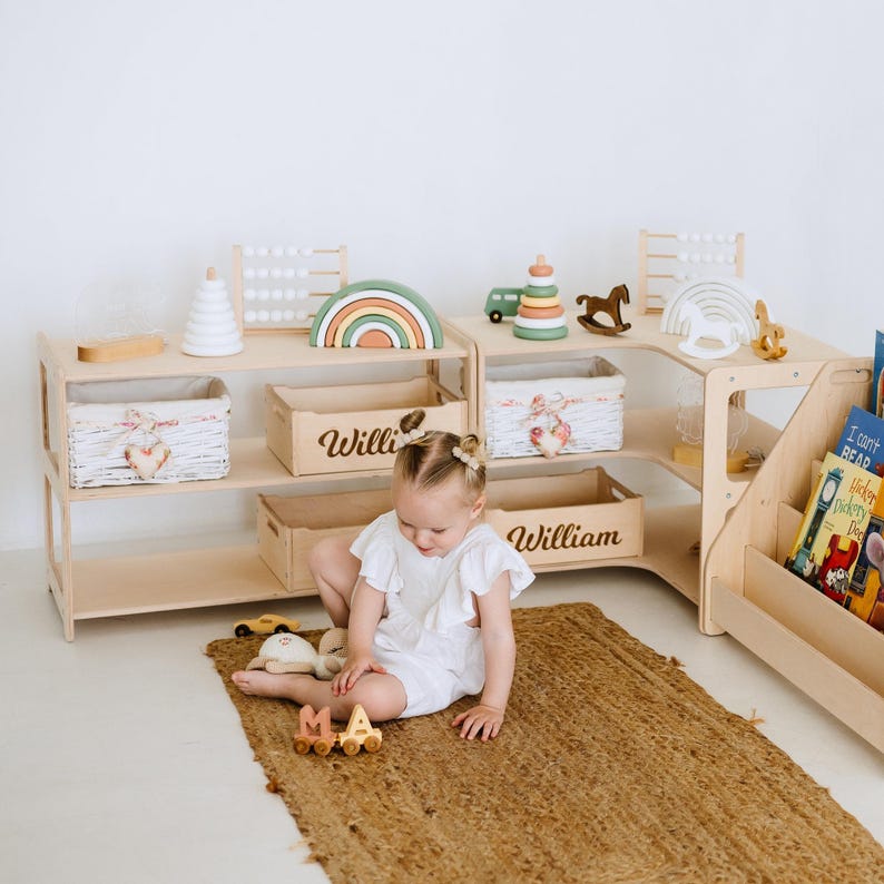 May include: A wooden corner shelf unit with three shelves on each side. The shelves are filled with toys, books, and baskets. The unit is painted white and has a natural wood finish. The shelves are labeled "Willie" and "William". A child is playing with toys on the floor in front of the shelves.
