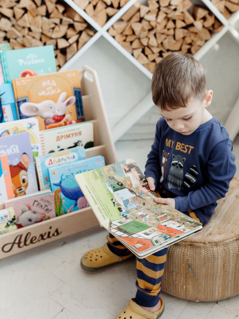 Pu&ograve; includere: Un bambino piccolo &egrave; seduto su un cuscino intrecciato e legge un libro illustrato colorato. Indossa pantaloni a righe blu e gialle e una camicia blu con la scritta "My Best" stampata. Il libro &egrave; aperto a una pagina con illustrazioni di una scena di fattoria. Il bambino &egrave; seduto davanti a una libreria di legno piena di libri per bambini. La libreria &egrave; etichettata "Alexis".