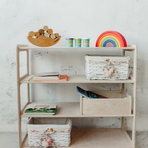 May include: A white wooden shelf unit with four shelves. The top shelf has a wooden rainbow toy, three small containers of playdough, and a wooden toy with stacked circles. The second shelf has a white wicker basket with a heart-shaped fabric accent. The third shelf has a stack of books. The fourth shelf has a white wicker basket with a heart-shaped fabric accent. The bottom shelf is empty.