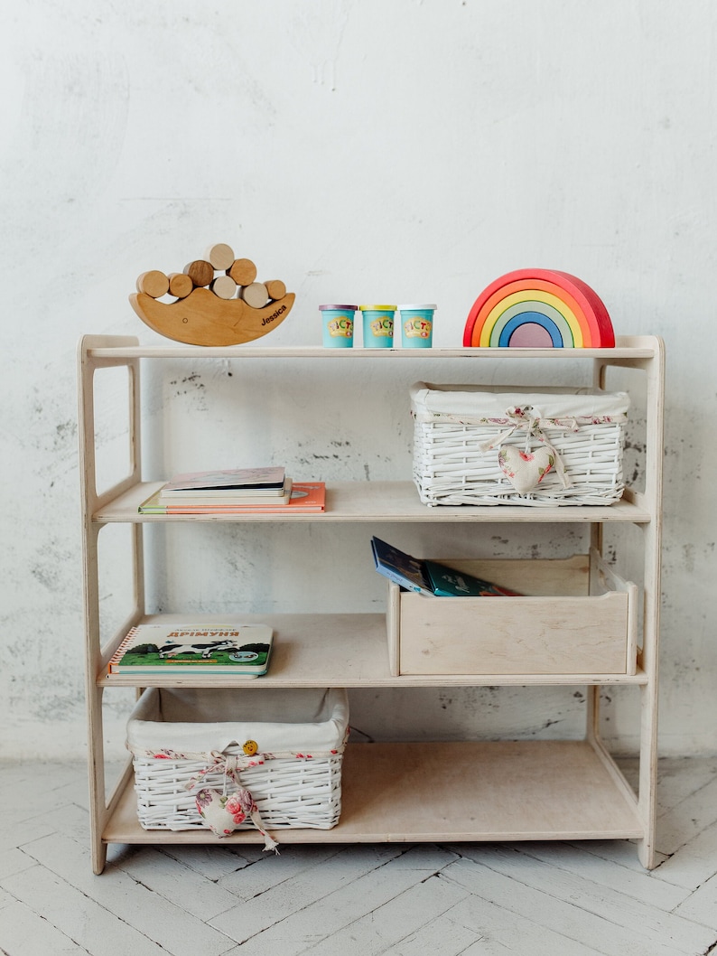 May include: A white wooden shelf unit with four shelves. The top shelf has a wooden rainbow toy, three small containers of playdough, and a wooden toy with stacked circles. The second shelf has a white wicker basket with a heart-shaped fabric accent. The third shelf has a stack of books. The fourth shelf has a white wicker basket with a heart-shaped fabric accent. The bottom shelf is empty.