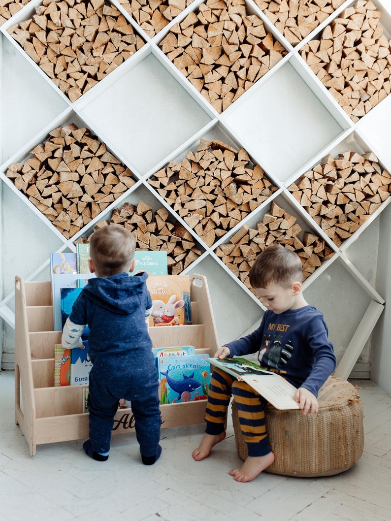 Pu&ograve; includere: Due bambini stanno leggendo libri. Un bambino &egrave; in piedi accanto a una libreria di legno piena di libri per bambini colorati. L'altro bambino &egrave; seduto su un cuscino intrecciato e sta leggendo un libro. La libreria ha il nome "Alex" scritto su di essa.