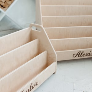 Pu&ograve; includere: Due librerie in legno con nomi personalizzati, "Taylor" e "Alexis", sono mostrate su un pavimento bianco. Le librerie sono in legno chiaro e hanno un design semplice.