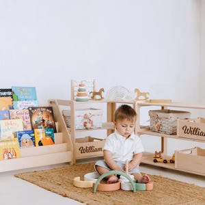 May include: A light wood bookshelf with a variety of children's books, a wooden toy with colorful rings, and a wooden rocking horse. A child is playing with a wooden stacking toy on a light brown rug. The bookshelf and toy storage units are personalized with the name "William".