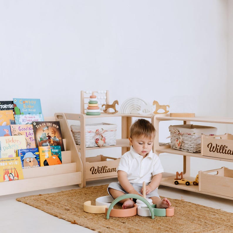 May include: A light wood bookshelf with a variety of children's books, a wooden toy with colorful rings, and a wooden rocking horse. A child is playing with a wooden stacking toy on a light brown rug. The bookshelf and toy storage units are personalized with the name "William".