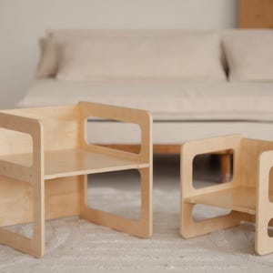 May include: Two small, light-colored wooden chairs with open rectangular cutouts on the sides. The chairs have a simple, modern design and are placed on a light-colored rug. The background includes a neutral-toned sofa.