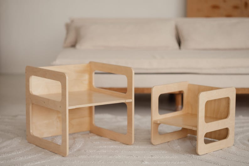 May include: Two small, light-colored wooden chairs with open rectangular cutouts on the sides. The chairs have a simple, modern design and are placed on a light-colored rug. The background includes a neutral-toned sofa.