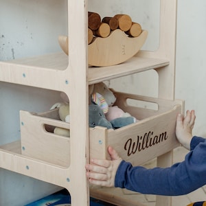 May include: A light wood bookshelf with four shelves and a drawer. The drawer is labeled "William". The bookshelf is filled with toys and books.
