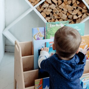 May include: A wooden bookshelf with multiple shelves filled with colorful children's books. A young child is reaching for a book on the top shelf.