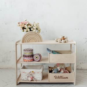 May include: A white wooden shelf unit with three shelves. The top shelf has a wooden rainbow toy and a wooden geometric toy. The middle shelf has three colorful storage containers. The bottom shelf has a white wicker basket and a wooden box with the name "William" engraved on it.