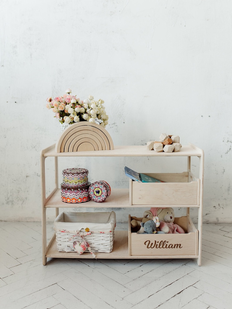 May include: A white wooden shelf unit with three shelves. The top shelf has a wooden rainbow toy and a wooden geometric toy. The middle shelf has three colorful storage containers. The bottom shelf has a white wicker basket and a wooden box with the name "William" engraved on it.