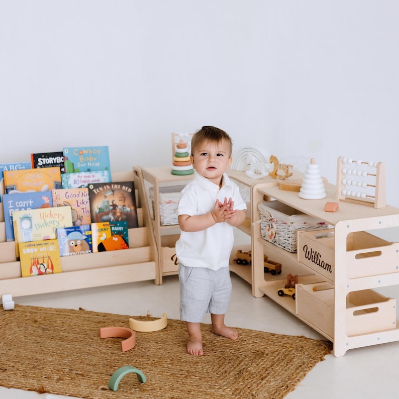 May include: A child stands near wooden bookshelves and storage units. The shelves display colorful children's books. The storage units have open shelves and drawers. A woven rug and wooden toys are on the floor. The name "William" is on a drawer.