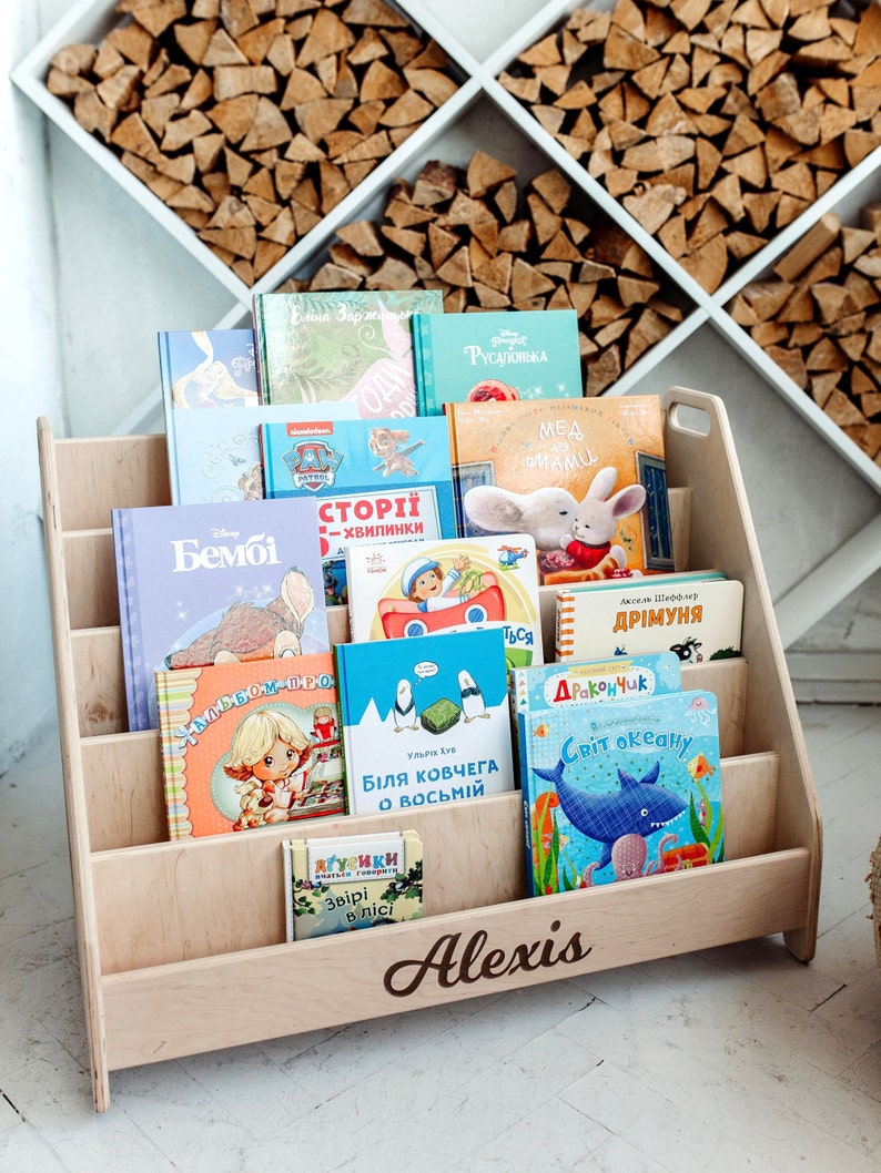 May include: A wooden bookshelf with five shelves, filled with colorful children's books. The bookshelf is light brown and has the name "Alexis" engraved on the bottom shelf.