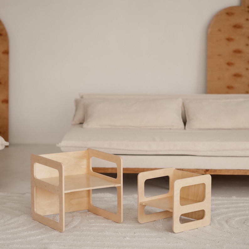 May include: Two small, light-colored wooden chairs with open sides and a shelf. The chairs are placed on a white rug in front of a neutral-toned sofa. The chairs have a simple, modern design.