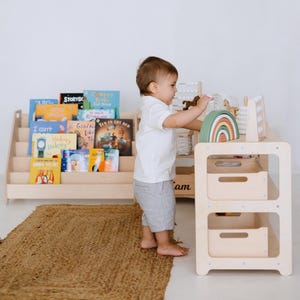 May include: A wooden children's play set with a rainbow, abacus, and storage drawers. A child is interacting with the toys. A wooden bookshelf is in the background, filled with colorful children's books. A woven rug is on the floor.