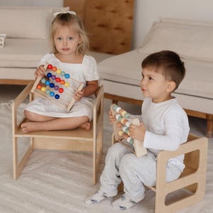 May include: Two children sit in wooden chairs, each holding a wooden abacus. One abacus has colorful beads, while the other has pastel-colored beads. The chairs and abacuses are made of light-colored wood, creating a playful and educational scene.