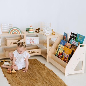 May include: A child's playroom with wooden furniture, including a bookshelf filled with colorful books, and a multi-level storage unit. The unit has wooden toy storage boxes labeled "Willi" and "William". Decorative wooden toys and a jute rug complete the scene.