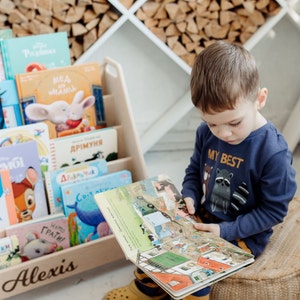 May include: A young boy sits on a woven cushion, reading a colorful book with illustrations of farm animals and a truck. He is wearing a blue shirt with the words "My Best" and a raccoon graphic. The book is open on his lap, and he is looking at the pages. The book is sitting on a wooden bookshelf with the name "Alexis" written on the front. The bookshelf is filled with other books, some of which are in Russian.