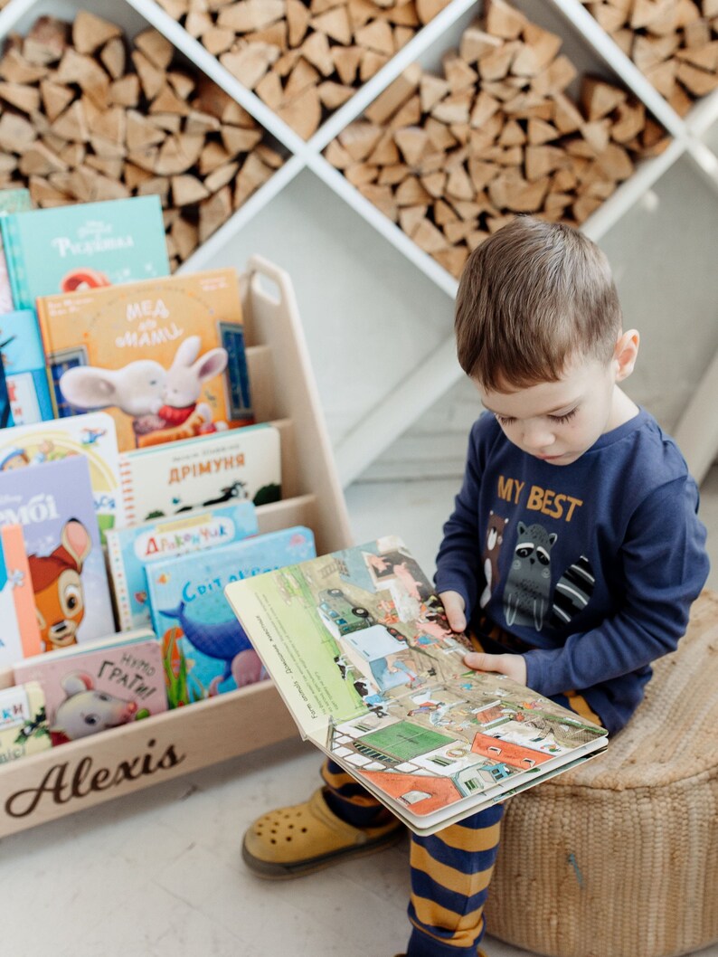 May include: A young boy sits on a woven cushion, reading a colorful book with illustrations of farm animals and a truck. He is wearing a blue shirt with the words "My Best" and a raccoon graphic. The book is open on his lap, and he is looking at the pages. The book is sitting on a wooden bookshelf with the name "Alexis" written on the front. The bookshelf is filled with other books, some of which are in Russian.