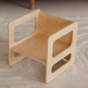 May include: A natural wood step stool with a square design. The stool has a small shelf and two rectangular cutouts on each side for handles. The wood has a light, natural finish, and the stool is designed for children.