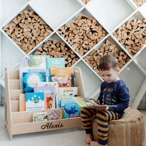 May include: A wooden bookshelf with a personalized name "Alexis" on the front. The bookshelf is filled with colorful children's books. A young boy is sitting on a woven cushion, reading a book. The bookshelf is in front of a wall with a decorative wood pattern.
