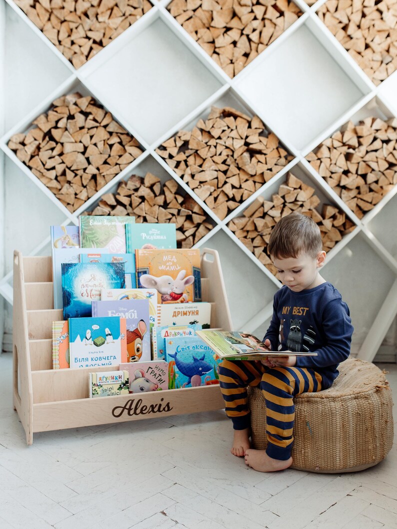 May include: A wooden bookshelf with a personalized name "Alexis" on the front. The bookshelf is filled with colorful children's books. A young boy is sitting on a woven cushion, reading a book. The bookshelf is in front of a wall with a decorative wood pattern.